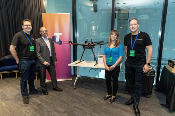 Two Telstra and two UoM staff members stand in front of the Telstra banner and drone at the 2023 FEIT Hackathon