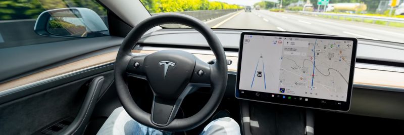 A car driving down a highway while the driver sits back with folded arms. A screen and Tesla logo are visible.