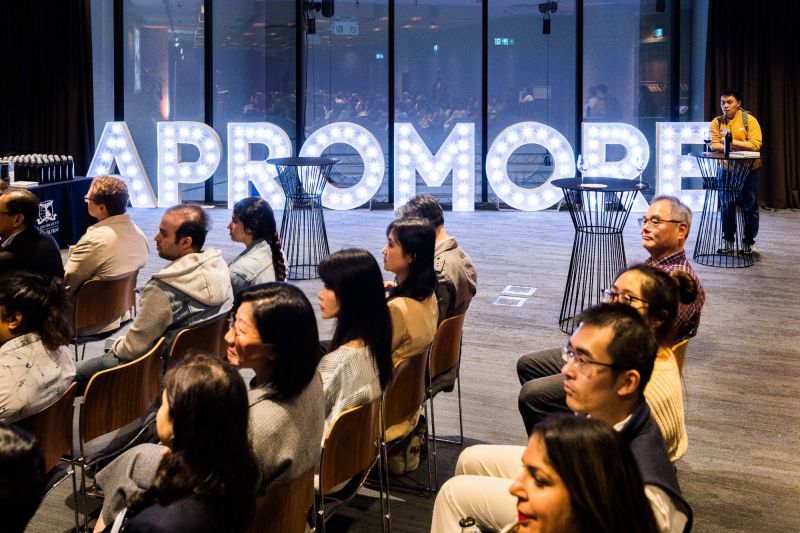 Audience seated next to bright 'Apromore' sign