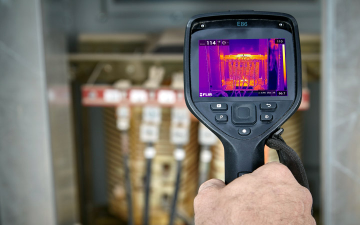 A hand holding a device showing a thermography heat map of electrical equipment