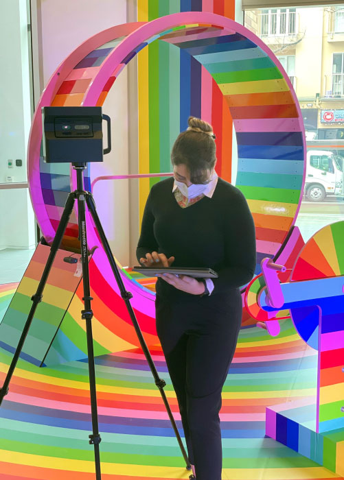 A woman dressed in black stands in front of rainbow stripped sculpture, working on a tablet