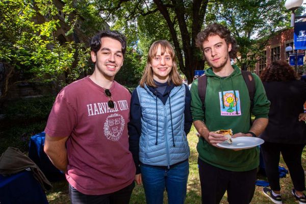 Three students stand in front of trees with one student holding a plate of food