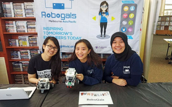 Three female students sitting in front of a laptop, with a Robogals banner in the background