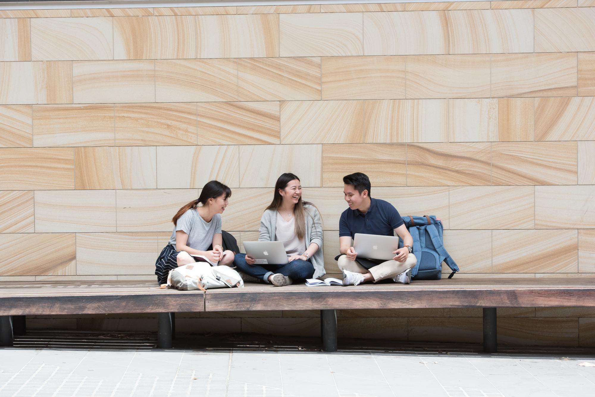 Three smiling students sit in front of a sandstone wall